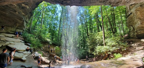 A scenic waterfall cascades over rocks, surrounded by lush green trees and people enjoying the natural beauty.