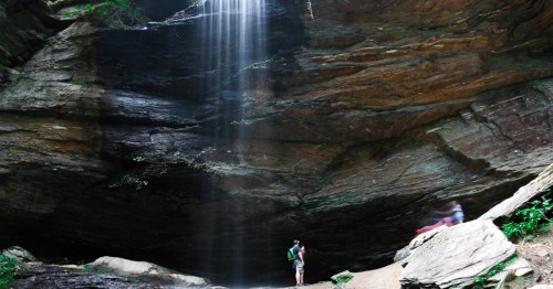 A person stands near a waterfall cascading down rocky cliffs in a lush, green forest setting.