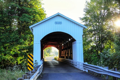 A white covered bridge with a wooden interior, surrounded by trees, leading to a sunlit road.