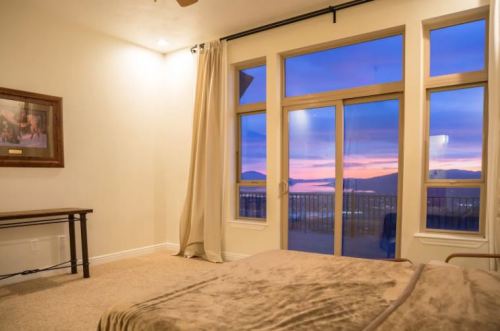 A cozy bedroom with a large window showcasing a colorful sunset over mountains. Soft lighting and neutral decor.