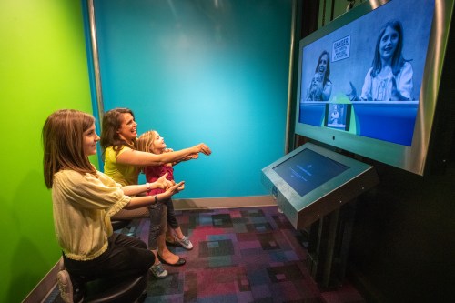 A family interacts with a touchscreen display, watching themselves on a large screen in a colorful room.