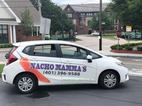 A white car with "Nacho Mammas" and a phone number on the side, parked on a street near a stop sign.