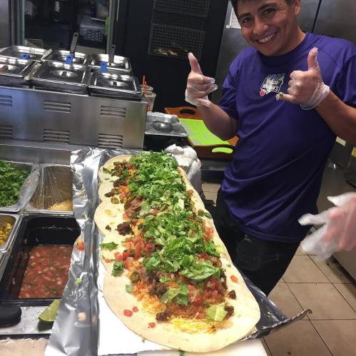 A chef gives a thumbs up next to a large burrito filled with meat, lettuce, and salsa, in a vibrant kitchen setting.