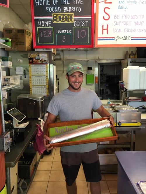 A smiling man holds a large burrito in a restaurant, with a scoreboard showing 123 for home and 10 for guest.