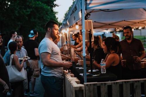 A lively outdoor market scene with people interacting at vendor booths under string lights during dusk.