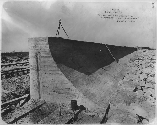 Black and white photo of a concrete sea wall under construction, with rocks and tracks in the background. Dated July 3, 1906.