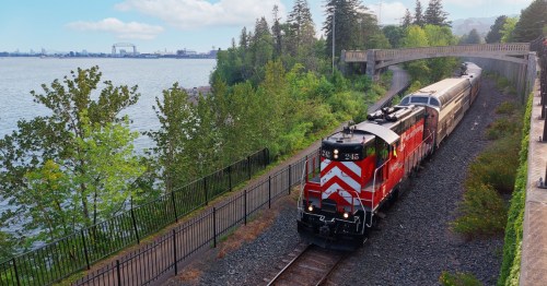 A red and white train travels along a scenic route by a lake, surrounded by greenery and a distant industrial area.