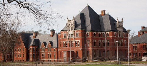 An old, abandoned brick building with a steep roof and ornate architectural details, surrounded by a fence and trees.