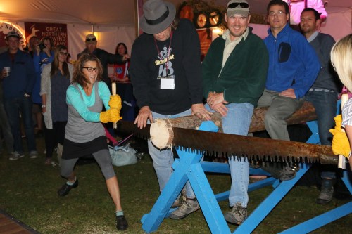 A woman saws a log while onlookers cheer at a lively outdoor event, showcasing a lumberjack competition.