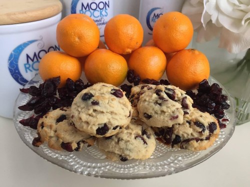 A plate of cookies surrounded by fresh oranges and dried cranberries, with decorative containers in the background.