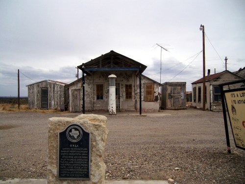 Abandoned building with a weathered facade, surrounded by small structures and a historical marker in a desolate landscape.