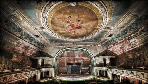 Abandoned theater interior with ornate ceiling mural, faded brick walls, and empty seating.
