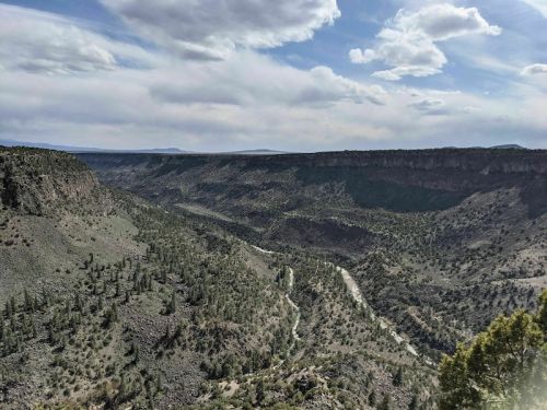 A panoramic view of a rugged canyon landscape with a winding river and scattered trees under a cloudy sky.