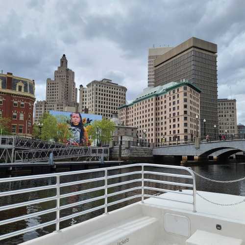 View of a city skyline with modern and historic buildings along a river, featuring a mural on one of the structures.