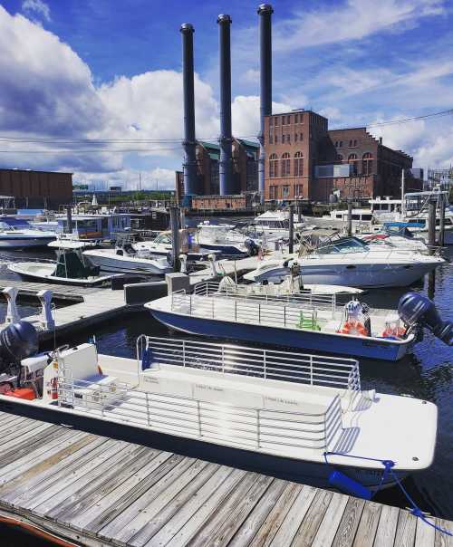 A marina with various boats docked, featuring a backdrop of industrial buildings and smokestacks under a blue sky.