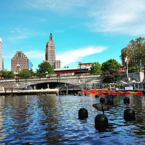 City skyline reflected in water, with buildings, trees, and a bridge under a blue sky. Kayaks are visible on the water.