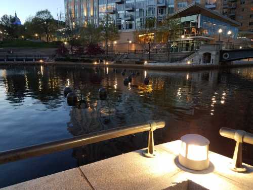 A serene waterfront scene at dusk, featuring lights reflecting on the water and modern buildings in the background.