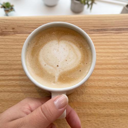 A hand holding a white cup of latte art with a heart design, set on a wooden surface.