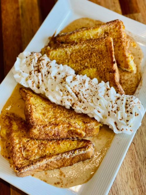 A plate of golden French toast topped with whipped cream and a dusting of cinnamon, served with a sweet sauce.