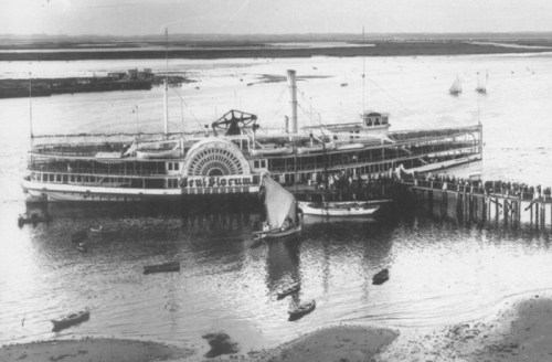 Historic black-and-white photo of a large steamboat docked at a pier, with people gathered on the deck and shore.