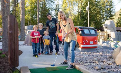 A woman putts on a mini-golf course while three children and a man watch, surrounded by trees and playful decor.