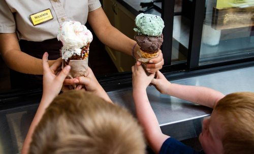 A server hands two ice cream cones to children at a counter, with colorful scoops of ice cream on top.