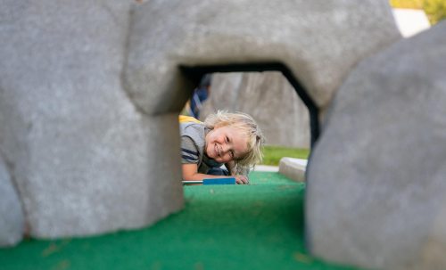 A child peeks through a stone archway while playing on a mini-golf course, smiling at the camera.