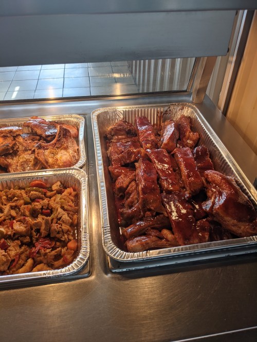 A tray of glazed barbecue ribs alongside another tray of mixed grilled meats and vegetables.