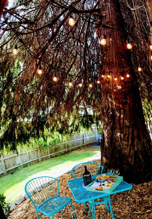 A cozy outdoor setting with a blue table and chairs under a tree, adorned with string lights and a spread of snacks.