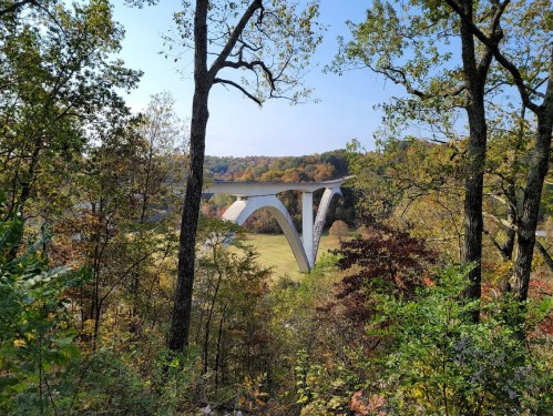 A modern bridge arches over a valley, surrounded by colorful autumn trees and clear blue skies.