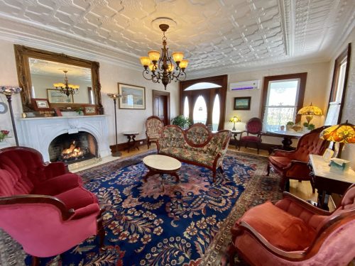 A cozy living room with ornate decor, a fireplace, elegant furniture, and a patterned rug under a chandelier.
