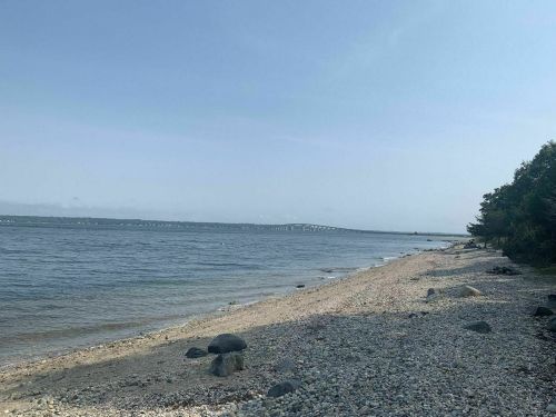 A serene beach scene with pebbles, calm water, and a distant bridge under a clear sky.