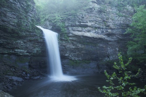 A serene waterfall cascades over rocky cliffs into a calm pool, surrounded by lush greenery and misty atmosphere.