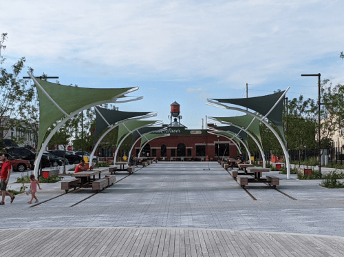A spacious outdoor area with green canopies, benches, and a building in the background, under a partly cloudy sky.