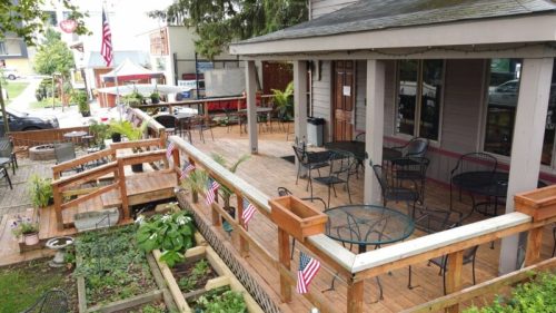 Outdoor café with wooden deck, tables, and chairs, decorated with small American flags and surrounded by greenery.