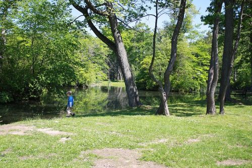 A child walks along a grassy path by a serene pond surrounded by lush green trees.