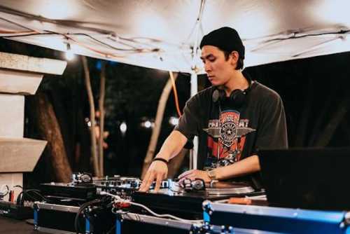 A DJ wearing a black cap and t-shirt mixes music at an outdoor event under a tent with string lights.