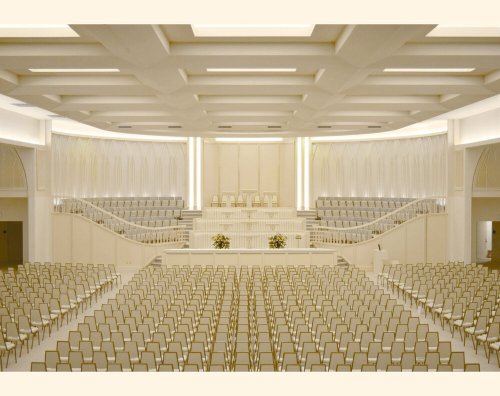 A spacious, modern auditorium with rows of empty chairs and elegant white decor, featuring a stage at the front.