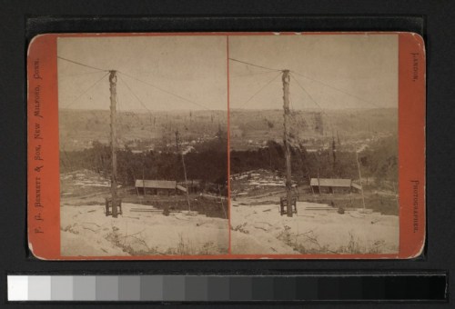 A vintage stereoscopic image showing a landscape with power lines and a small building near a rocky terrain.