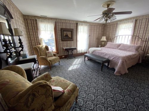 Cozy bedroom with a large bed, armchair, and patterned wallpaper, featuring natural light from windows.