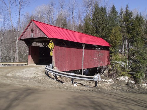 A red-roofed covered bridge sits at a curve in a dirt road, surrounded by trees and a guardrail.