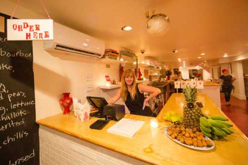 A smiling woman stands behind a counter with a cash register, surrounded by fresh fruits and a sign that says "Order Here."