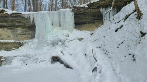 A frozen waterfall surrounded by snow-covered rocks and trees in a winter landscape.