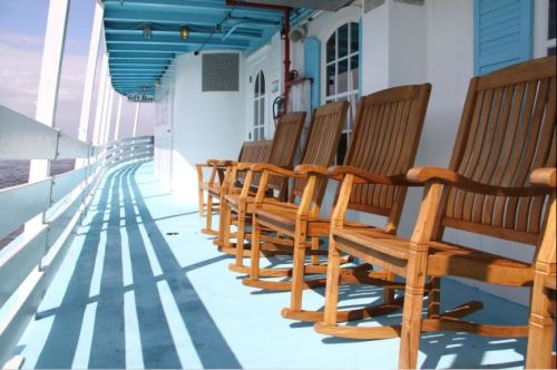 A row of wooden rocking chairs on a blue deck, with sunlight casting shadows on the floor.
