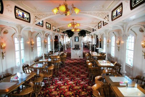 Elegant restaurant interior with ornate decor, wooden chairs, and stained glass windows, featuring a rich carpet design.