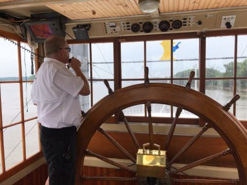 A person in a white shirt stands at the helm of a boat, looking out through large windows.