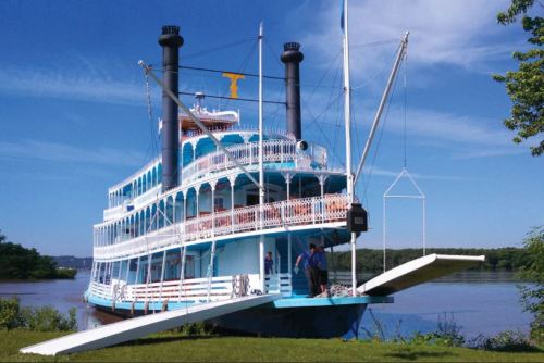 A large blue and white riverboat with three smokestacks docked by the shore, with people boarding from a ramp.
