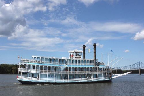 A large paddleboat cruises on a river under a blue sky with clouds, with a bridge visible in the background.