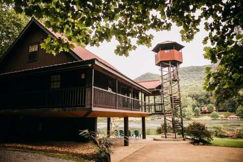 A rustic cabin with a tower, surrounded by trees and a river, set against a backdrop of rolling hills.