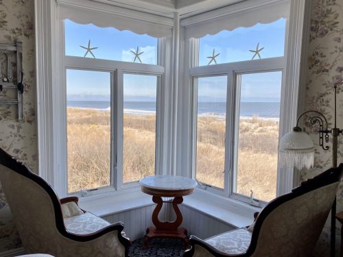 Cozy seating area with two chairs and a small table, overlooking a beach and ocean through large bay windows.